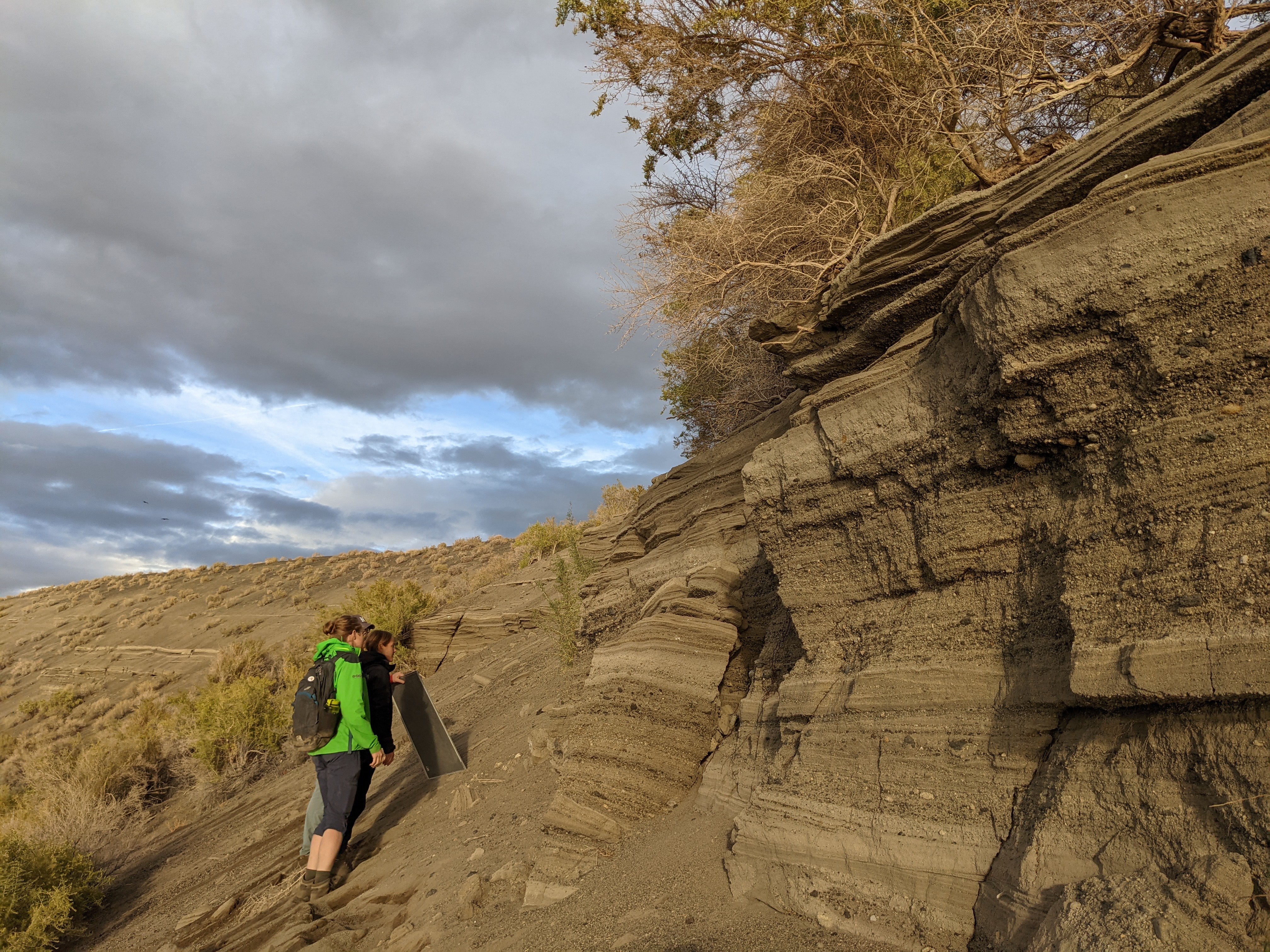 Two people examine tilted layers of volcanic ash and sediment near Soda Lake, Nevada. One holds a sample board. The outcrop is steep and textured, with dry shrubs and a cloudy sky in the background.